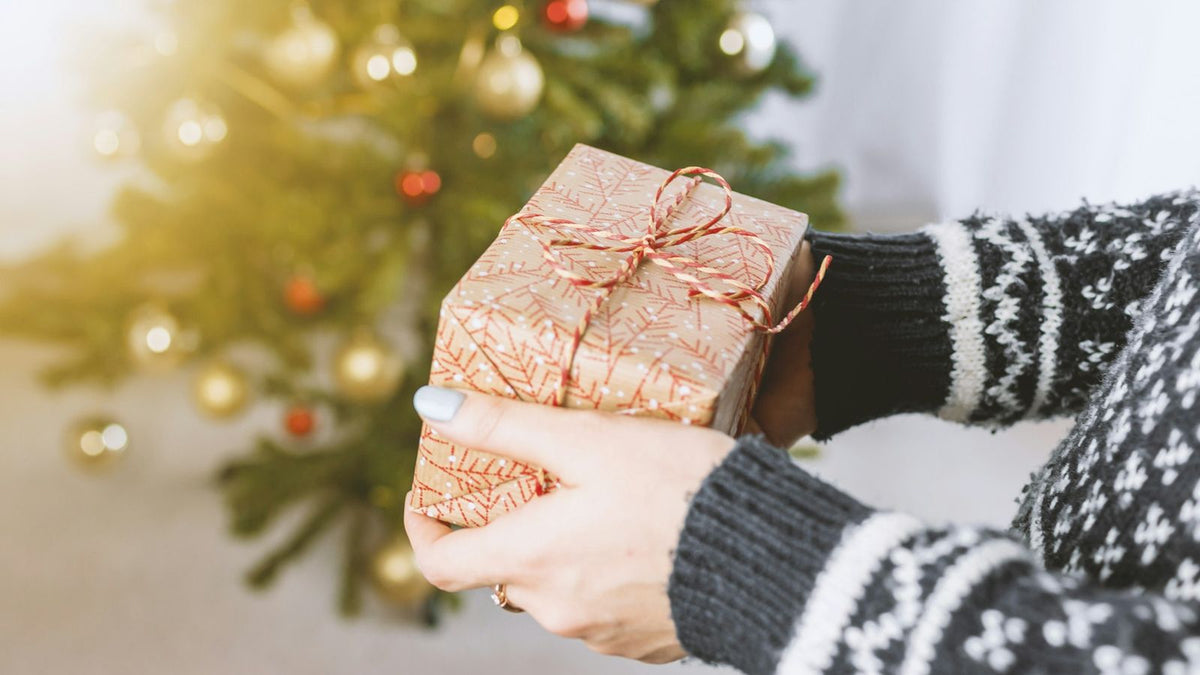 飾りを付けたクリスマスツリーの前で、赤い柄の包装紙と赤白のひもで包んだ小さな箱を両手で持つ、ノルディック柄のニットの手元