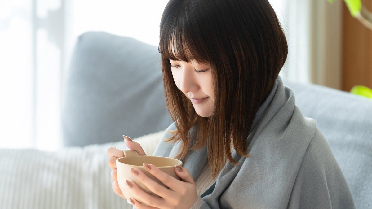 肩から水色のブランケットを掛けた女性が、ソファの前で両手でカップを包むように持っている