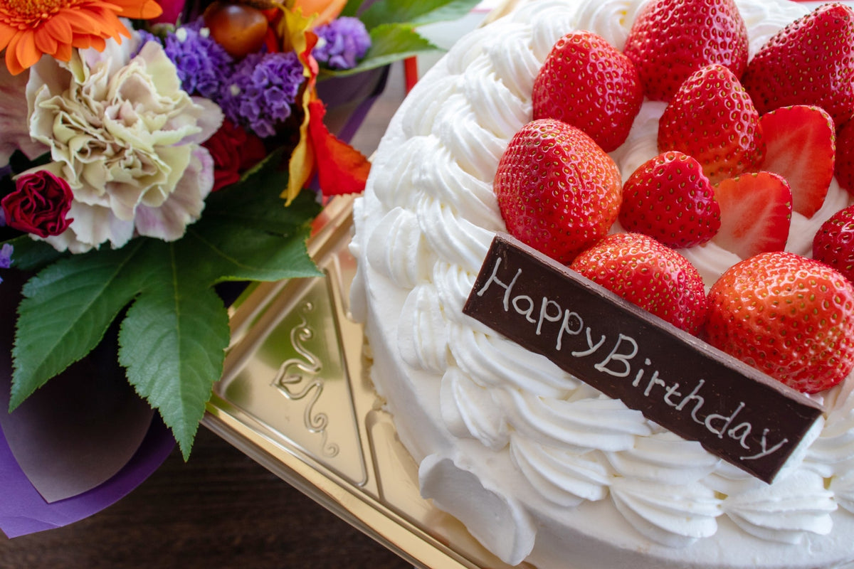 いちごを並べた白いホールケーキに「Happy Birthday」の飾りがのり、そばに花束が置かれている