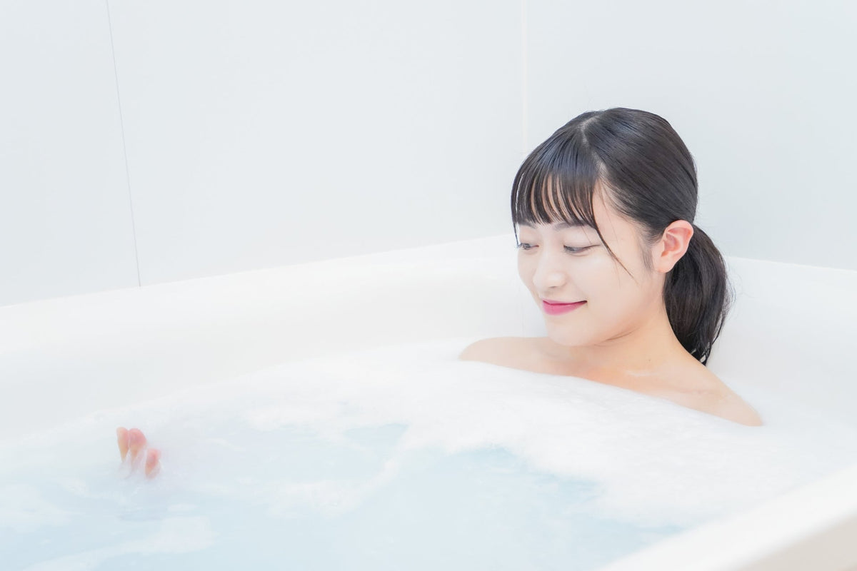 白い浴槽で泡の立った湯につかり、目を閉じてほほえむ髪を結んだ女性