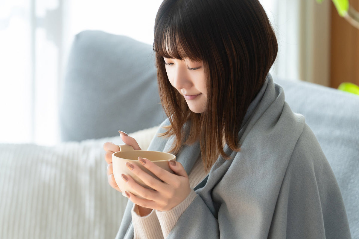 肩から水色のブランケットを掛けた女性が、ソファの前で両手でカップを包むように持っている