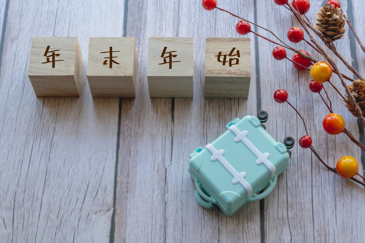 「年末年始」と書かれた4つの木のブロックと、水色のミニチュアのスーツケース、赤い実と松ぼっくりの枝が木目の台に並んでいる