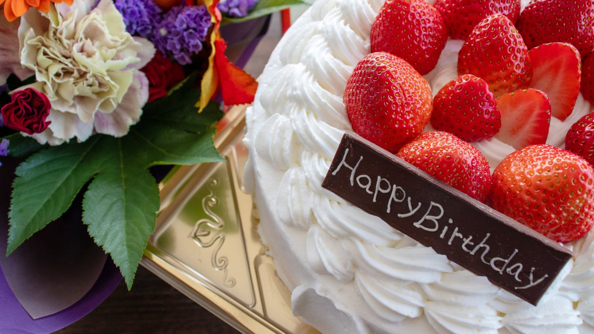 いちごを並べた白いホールケーキに「Happy Birthday」の飾りがのり、そばに花束が置かれている