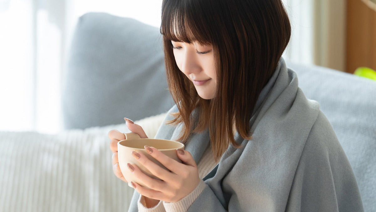 灰色のブランケットを肩から掛け、ソファの前でカップを両手で持つ女性