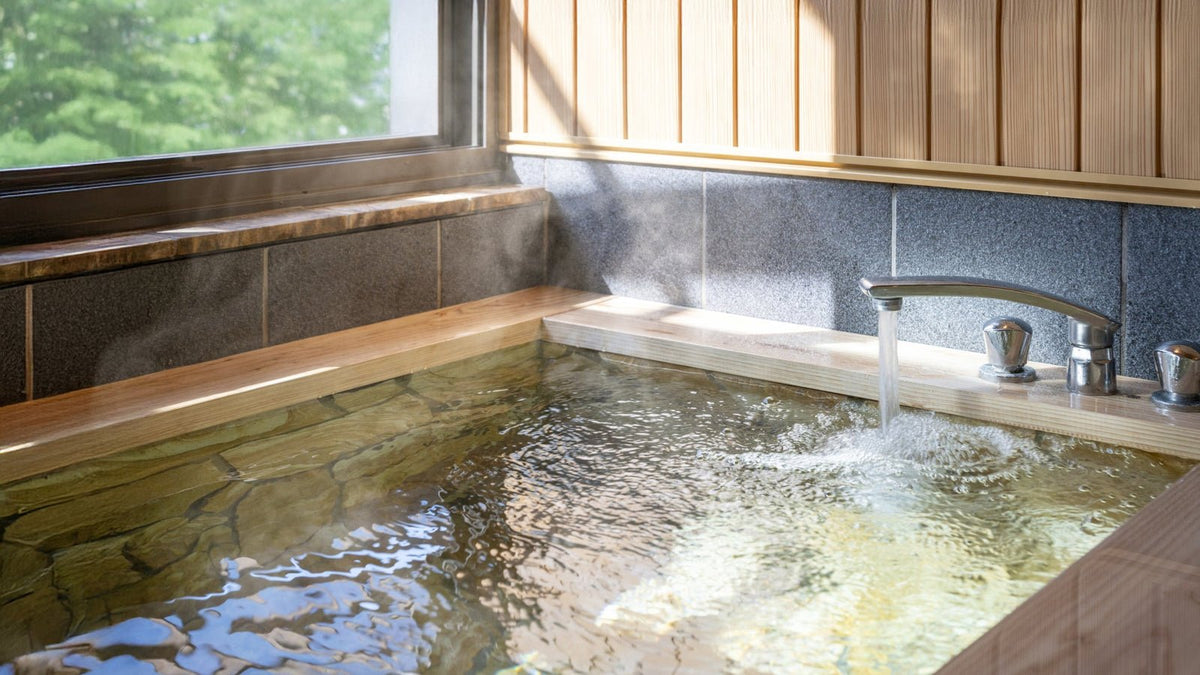 窓から日が差す木の縁の浴槽に、蛇口からお湯が注がれ、湯気が立っている