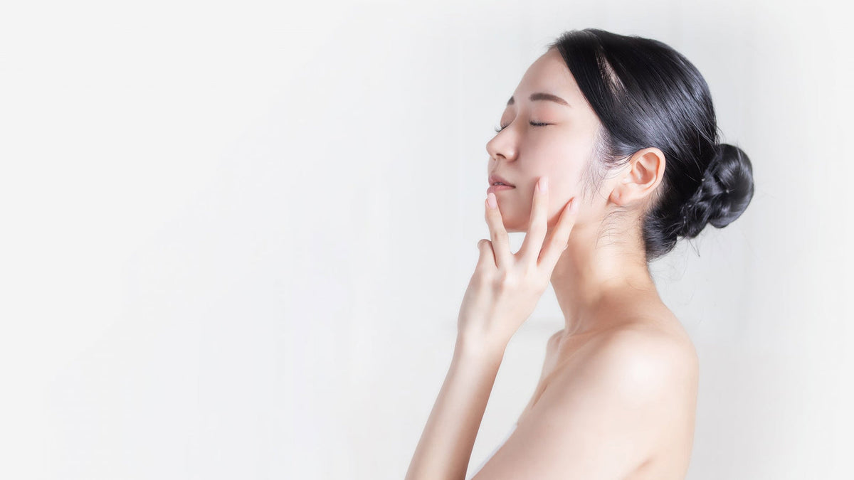 白い背景の前で、目を閉じて頬に指先を添える、髪をまとめた女性の横顔