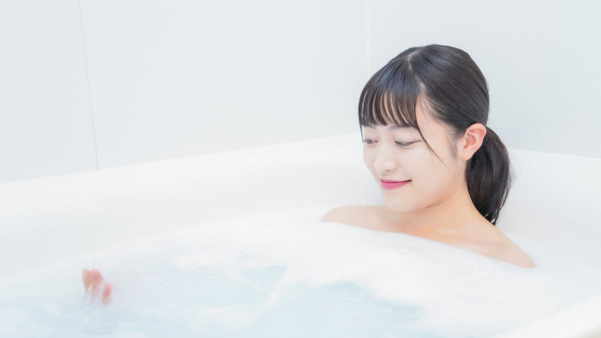 白い浴槽で泡の立った湯につかり、目を閉じてほほえむ髪を結んだ女性