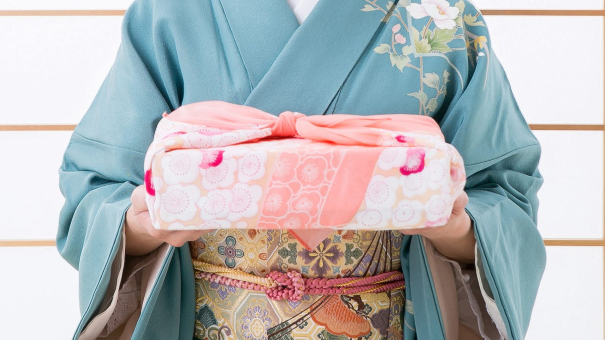 障子の前で、青緑の着物の人が桃色の梅の柄の風呂敷で包んだ箱を両手で差し出す手元（顔は写っていない）