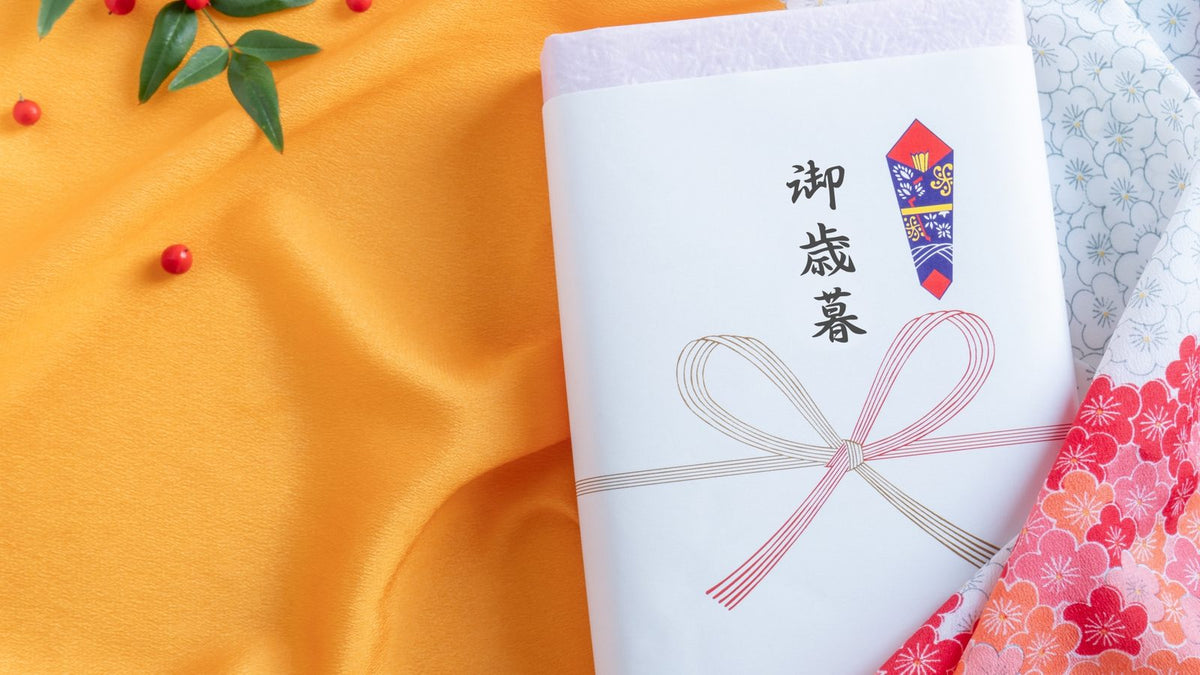 「御歳暮」と書いた紅白の蝶結びの水引ののし紙を掛けた箱と、山吹色のちりめんの布、梅の柄の布、南天の実と葉