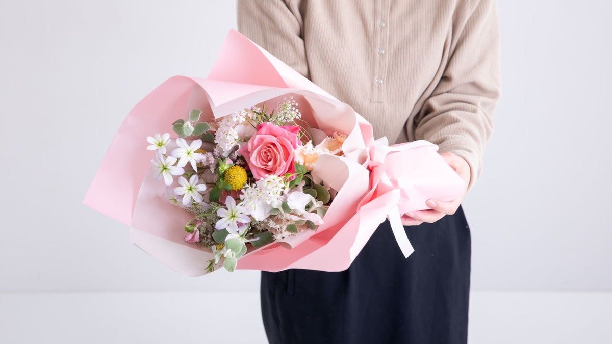 ピンクの包装紙で包んだ、ピンクのバラや白い小花の花束を両手で抱えるベージュのカーディガンの人（顔は写っていない）